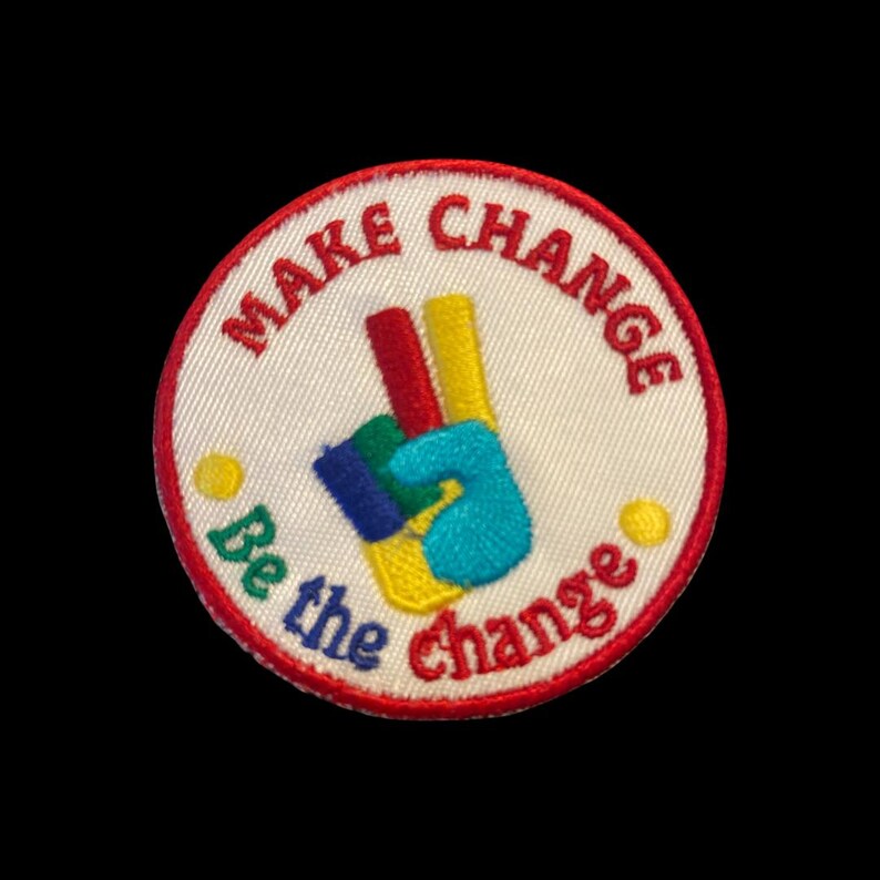 K&ouml;nnte beinhalten: Gestickter Aufn&auml;her mit wei&szlig;em Hintergrund, rotem Rand und den Worten "MAKE CHANGE" und "Be the change" in Rot und Gr&uuml;n. Der Aufn&auml;her zeigt ein farbenfrohes Peace-Zeichen mit roten, gelben, blauen und t&uuml;rkisfarbenen Fingern.