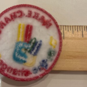 K&ouml;nnte beinhalten: Ein runder, gestickter Aufn&auml;her mit rotem Rand und den Worten "MAKE CHANGE HAPPEN" in einem kreisf&ouml;rmigen Muster. Der Aufn&auml;her zeigt ein farbenfrohes Handsymbol mit blauen, gelben, roten und gr&uuml;nen Details. Ein Holzlineal liegt neben dem Aufn&auml;her.