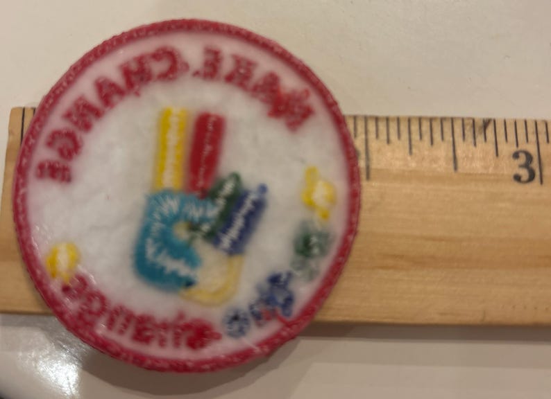 K&ouml;nnte beinhalten: Ein runder, gestickter Aufn&auml;her mit rotem Rand und den Worten "MAKE CHANGE HAPPEN" in einem kreisf&ouml;rmigen Muster. Der Aufn&auml;her zeigt ein farbenfrohes Handsymbol mit blauen, gelben, roten und gr&uuml;nen Details. Ein Holzlineal liegt neben dem Aufn&auml;her.