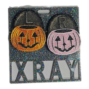 Puede incluir: Dos porta-películas de rayos X con temática de Halloween con fondo de purpurina negra. Los porta-películas tienen forma de calabaza con un sombrero negro en la parte superior. Una calabaza es naranja y la otra es rosa. Los porta-películas están etiquetados con "L" y "R" para izquierda y derecha. La palabra "XRAY" está impresa en negro en la parte inferior de los porta-películas.