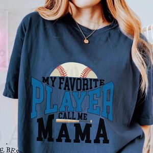 Puede incluir: Una camiseta azul marino con el texto "My Favorite Player Call Me Mama" en letras blancas y negras. Un gráfico de béisbol y bate está por encima del texto.
