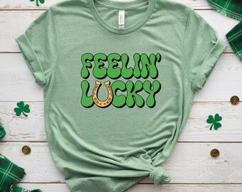 May include: A light green t-shirt with the words "FEELIN' LUCKY" in green bubble letters and a gold horseshoe graphic. The shirt is a crew neck style and is knotted at the bottom. Shamrock and gold coin decorations are scattered around the shirt.