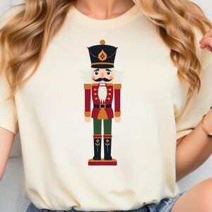 May include: Cream-colored t-shirt featuring a colorful illustration of a nutcracker soldier. The nutcracker has a black hat, a white mustache, and is dressed in a red and green uniform. The design is centered on the shirt.