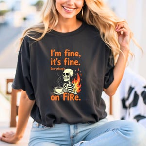 Może przedstawiać: Czarny T-shirt z nadrukiem szkieletu trzymającego kubek kawy i płomienia za nim. Tekst na koszulce brzmi "I'm fine, it's fine. Everything's on Fire."