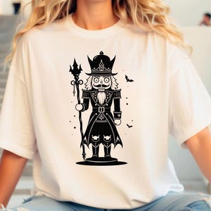 May include: White t-shirt featuring a black and white illustration of a nutcracker soldier holding a staff. The nutcracker has a mustache, beard, and a decorative hat. Small bat silhouettes are scattered around the design. A festive graphic tee.