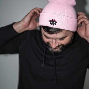 Puede incluir: Un gorro rosa claro con un diseño de ojo bordado en negro y rojo. El gorro está hecho de un material de punto suave. La persona de la imagen lleva una sudadera con capucha negra.