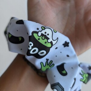 Op de afbeelding: Een lichtblauwe scrunchie met een Halloween-thema. Het patroon bevat een ketel met een spook, het woord "BOO", sterren en groene en zwarte accenten. De scrunchie is om een pols gewikkeld.