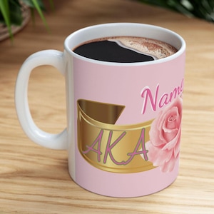 Puede incluir: Una taza de café de cerámica blanca con fondo rosa y un anillo dorado con la palabra "AKA" en letras rosas. La taza también tiene una rosa rosa y el texto "Name" en letras rosas.