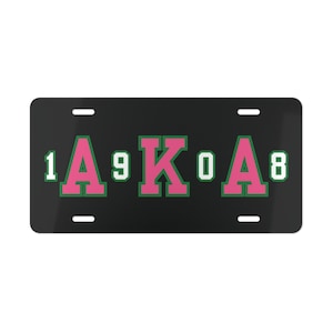 May include: Black license plate with white numbers and letters. The letters "AKA" are in pink with a green outline. The numbers "1908" are on either side of the letters.