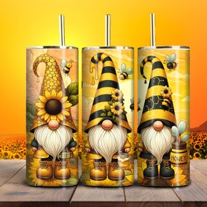May include: Three stainless steel tumblers with bee-themed gnome designs. Each tumbler features a different gnome with a long white beard, a pointed hat, and a bee-related design. The tumblers have gold straws and are set against a sunflower field backdrop.