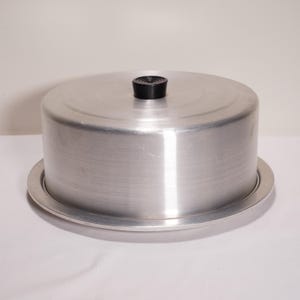 May include: A round, aluminum cake carrier with a lid and a black handle. The carrier sits on a matching base. The metal has a brushed finish. The cake carrier is designed to protect and transport baked goods.