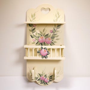 May include: A cream-colored wooden wall shelf featuring a heart-shaped cutout at the top, two tiers for storage, and two hooks at the bottom. The piece is decorated with hand-painted pink rose blossoms and green foliage accents throughout.