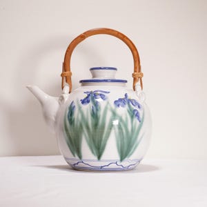 May include: White ceramic teapot with a bamboo handle and a lid. The teapot is decorated with blue iris flowers and green leaves. The handle is made of woven bamboo. The teapot has a spout for pouring.