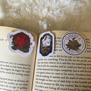 May include: A close-up of a book with two bookmarks. The bookmarks are both rectangular and feature illustrations. The first bookmark features a red door with a green vine and a red roof. The second bookmark features a white horse with a brown mane and tail. The text on the bookmark reads "Prancing Pony". The text on the book reads "Gamgee Potato Farm".