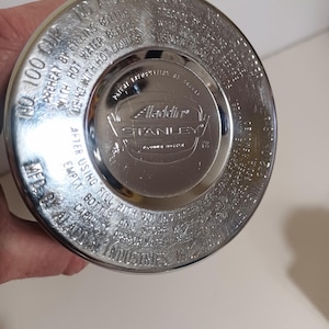 May include: A close-up of the bottom of a silver-colored Aladdin Stanley thermos. The lid has embossed text including "No. 100 Cup" and "1 Quart". The Aladdin Stanley logo is in the center.