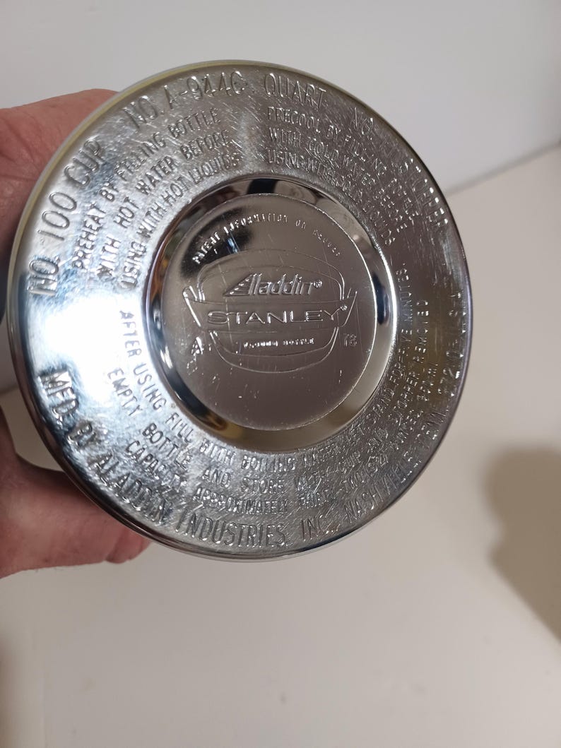 May include: A close-up of the bottom of a silver-colored Aladdin Stanley thermos. The lid has embossed text including "No. 100 Cup" and "1 Quart". The Aladdin Stanley logo is in the center.