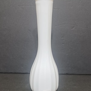 May include: A tall, white milk glass bud vase with a slender neck and a bulbous, vertically ribbed base. The rim features a subtle scalloped edge. A small square wooden tile with the letter F and the number 4 is placed at the base of the vase.