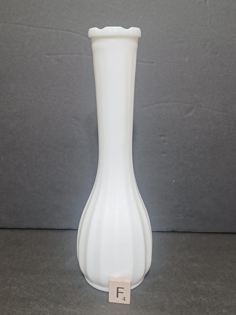 May include: A tall, white milk glass bud vase with a slender neck and a bulbous, vertically ribbed base. The rim features a subtle scalloped edge. A small square wooden tile with the letter F and the number 4 is placed at the base of the vase.