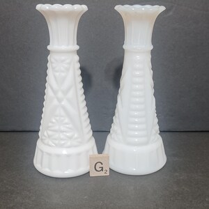 May include: Two white milk glass candle holders with a flared rim and a tapered body. The left holder features a geometric diamond and star pattern, while the right holder has a vertical ribbed design. A small wooden tile with the text G2 is present.
