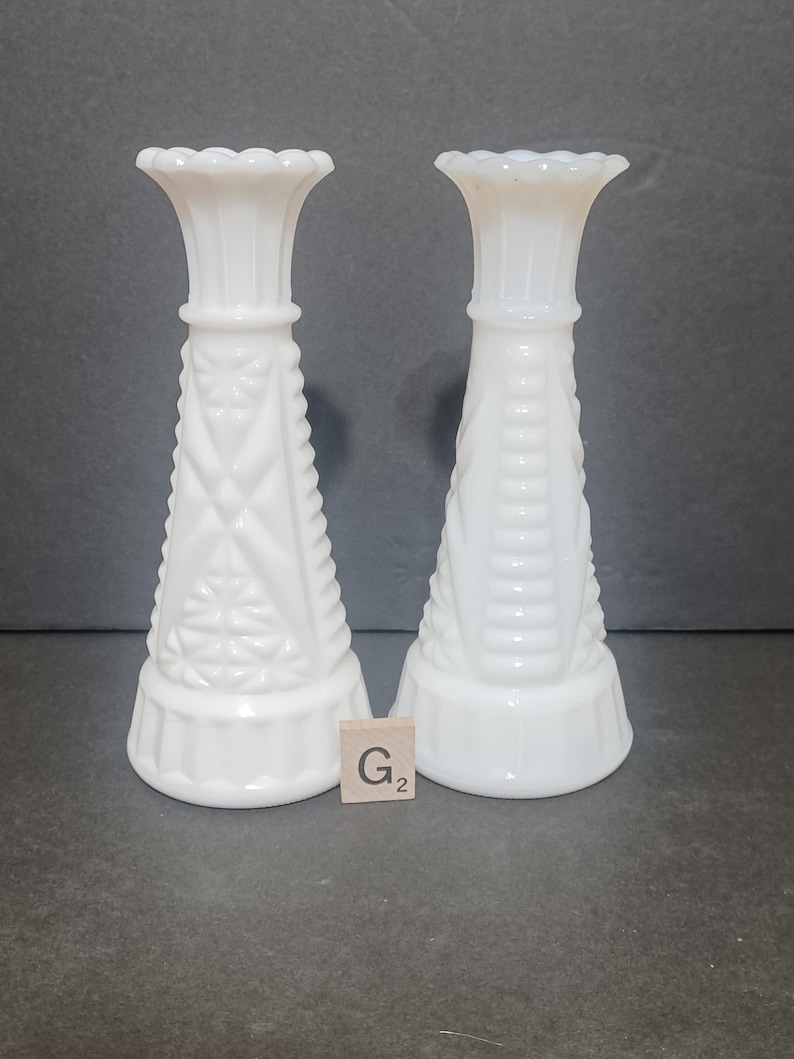 May include: Two white milk glass candle holders with a flared rim and a tapered body. The left holder features a geometric diamond and star pattern, while the right holder has a vertical ribbed design. A small wooden tile with the text G2 is present.