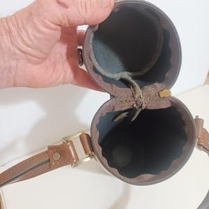 May include: A brown cylindrical case with a zipper, a brown leather strap, and a gold-colored buckle. The interior is lined with dark blue fabric. The case is open, revealing the interior.