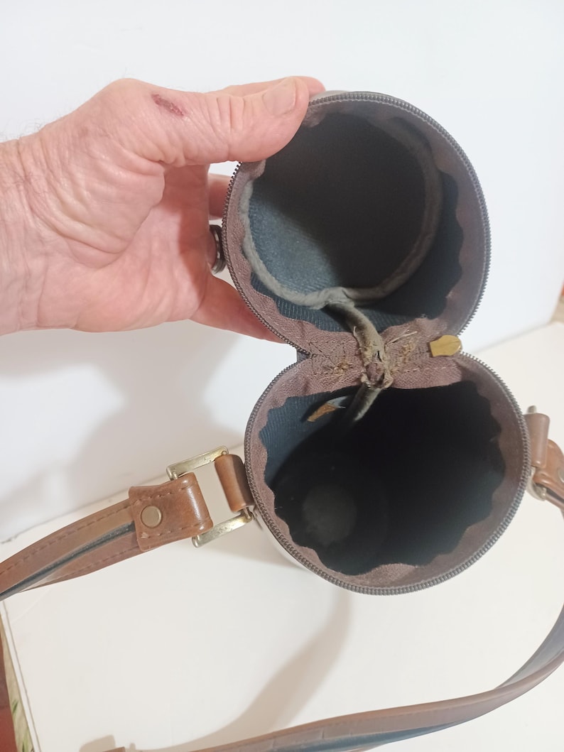 May include: A brown cylindrical case with a zipper, a brown leather strap, and a gold-colored buckle. The interior is lined with dark blue fabric. The case is open, revealing the interior.