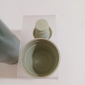 May include: An overhead view of a light green travel mug with a matching lid. The mug is open, revealing the interior. The lid is detached and placed next to the mug. The mug has a textured exterior and is set against a white background.