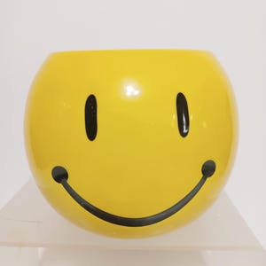May include: A bright yellow, spherical ceramic planter or bowl featuring a classic black smiley face design with two oval eyes and a wide, curved mouth. The surface is smooth and glossy, showcasing a vibrant yellow finish with bold black details.