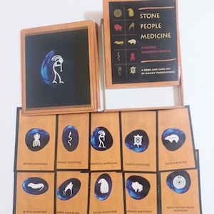 May include: A Native American oracle card set titled "Stone People Medicine" with a book and cards. The cards feature illustrations of animals and symbols, including a spider, snake, and eagle. The set is by Manny Twofeathers.