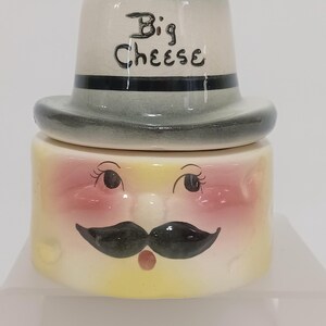 May include: A ceramic jar with a hat-shaped lid. The jar is yellow with a face design, including eyes, a mustache, and a red nose. The lid is light gray with the words "Big Cheese" in black.