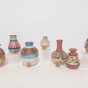 May include: A collection of small, decorative ceramic vases with various shapes and designs. The vases feature colors like red, blue, brown, and green, with geometric and abstract patterns. Each vase is displayed on a white surface.