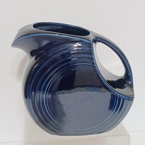 May include: A deep navy blue ceramic pitcher featuring a rounded body with concentric circular ring detailing on the side. The pitcher has a wide pouring spout and a curved handle integrated into the upper portion of the vessel's design.