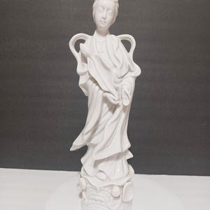 May include: A white porcelain figurine of a woman in flowing robes, with an elaborate hairstyle. The figure has a serene expression and is standing on a decorative base. The statue is approximately 12 inches tall.