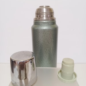 May include: A vintage green and silver thermos with a stainless steel lid and cup. The thermos has a textured finish and a silver cup with horizontal lines. The lid and cup are separate from the thermos.