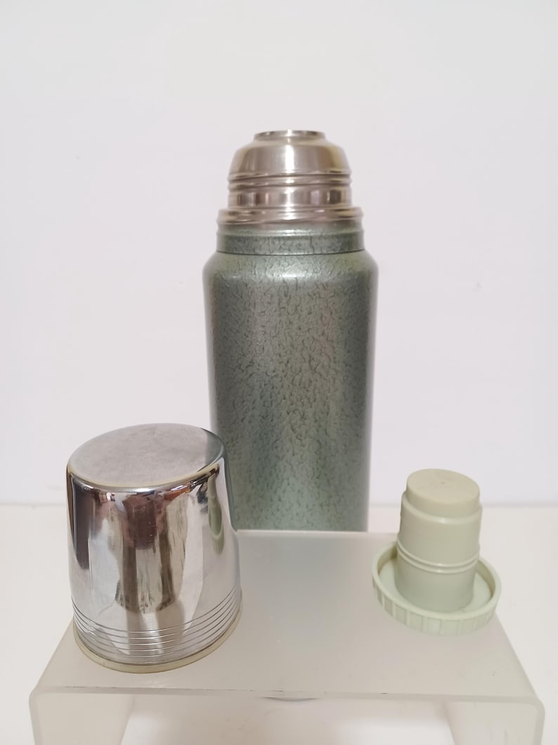May include: A vintage green and silver thermos with a stainless steel lid and cup. The thermos has a textured finish and a silver cup with horizontal lines. The lid and cup are separate from the thermos.