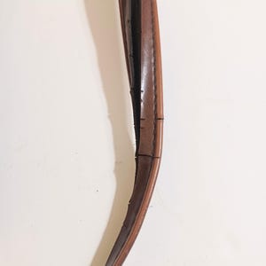 May include: Close-up of a brown leather handle, possibly from a bag or purse. The handle is curved and shows signs of wear, with visible cracks and imperfections along its length. The leather has a smooth, polished appearance.