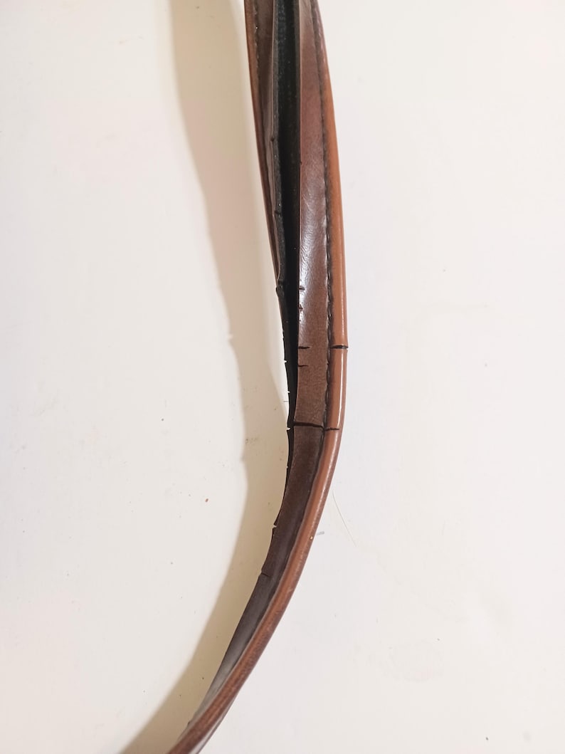 May include: Close-up of a brown leather handle, possibly from a bag or purse. The handle is curved and shows signs of wear, with visible cracks and imperfections along its length. The leather has a smooth, polished appearance.