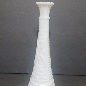 May include: A tall, white milk glass bud vase featuring a flared, scalloped rim and a tapered neck. The body of the vase is decorated with a raised diamond pattern and vertical ribbed detailing at the base, accompanied by a small wooden tile labeled E1.
