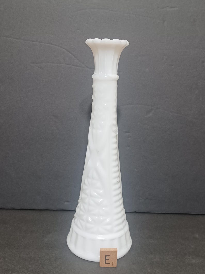 May include: A tall, white milk glass bud vase featuring a flared, scalloped rim and a tapered neck. The body of the vase is decorated with a raised diamond pattern and vertical ribbed detailing at the base, accompanied by a small wooden tile labeled E1.