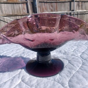 May include: A vintage, amethyst-colored glass bowl with a ruffled rim and a pedestal base. The bowl has a textured surface with small air bubbles. The base is a darker shade of purple.