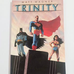 May include: A comic book titled "Trinity" by Matt Wagner, featuring Batman, Superman, and Wonder Woman. The cover art depicts the three superheroes standing together against a colorful sky. The DC Comics logo is visible.