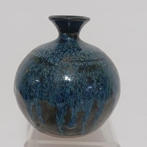 May include: A round, ceramic vase with a dark blue glaze that drips down the sides. The vase has a narrow neck and a glossy finish. The color transitions from dark blue to a lighter blue, creating a textured effect.