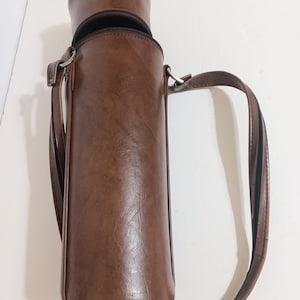 May include: A brown leather bottle carrier with a zipper closure and a shoulder strap. The cylindrical carrier is designed to hold a bottle and has a textured surface. The strap is attached with metal rings.