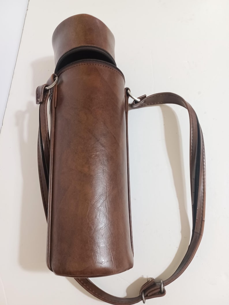 May include: A brown leather bottle carrier with a zipper closure and a shoulder strap. The cylindrical carrier is designed to hold a bottle and has a textured surface. The strap is attached with metal rings.