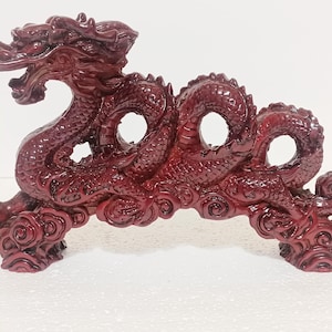 May include: A detailed, burgundy-colored dragon sculpture. The dragon is carved with intricate scales and features, including open circular designs in its body. The sculpture is mounted on a base with cloud-like details.
