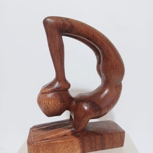 May include: A wooden sculpture of a person in a yoga pose. The figure is carved from a dark brown wood and is mounted on a rectangular base. The sculpture depicts a person bending backward, with one leg extended upwards.