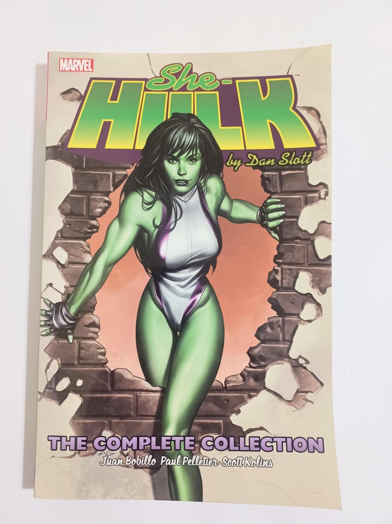 May include: A comic book cover for "She-Hulk" by Dan Slott. The cover features a green-skinned woman in a white and purple bodysuit breaking through a brick wall. The Marvel logo is visible at the top left, with the title in green and yellow.