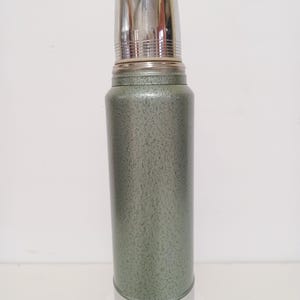 May include: A vintage green and silver thermos. The thermos has a textured green body with a silver lid and base. The lid is a cup-shaped design. The thermos is designed to keep liquids hot or cold.