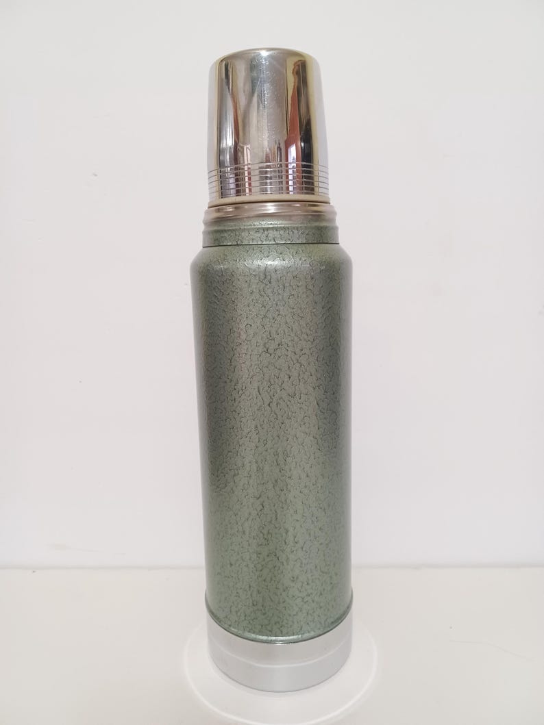 May include: A vintage green and silver thermos. The thermos has a textured green body with a silver lid and base. The lid is a cup-shaped design. The thermos is designed to keep liquids hot or cold.