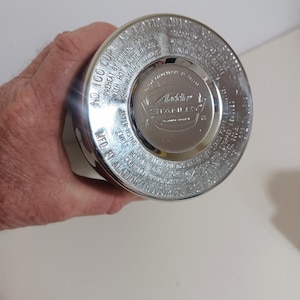 May include: A silver-colored Aladdin Stanley thermos cup. The lid is visible, with embossed text including "No. 100 Cup" and "Aladdin Stanley." The cup is held by a person.